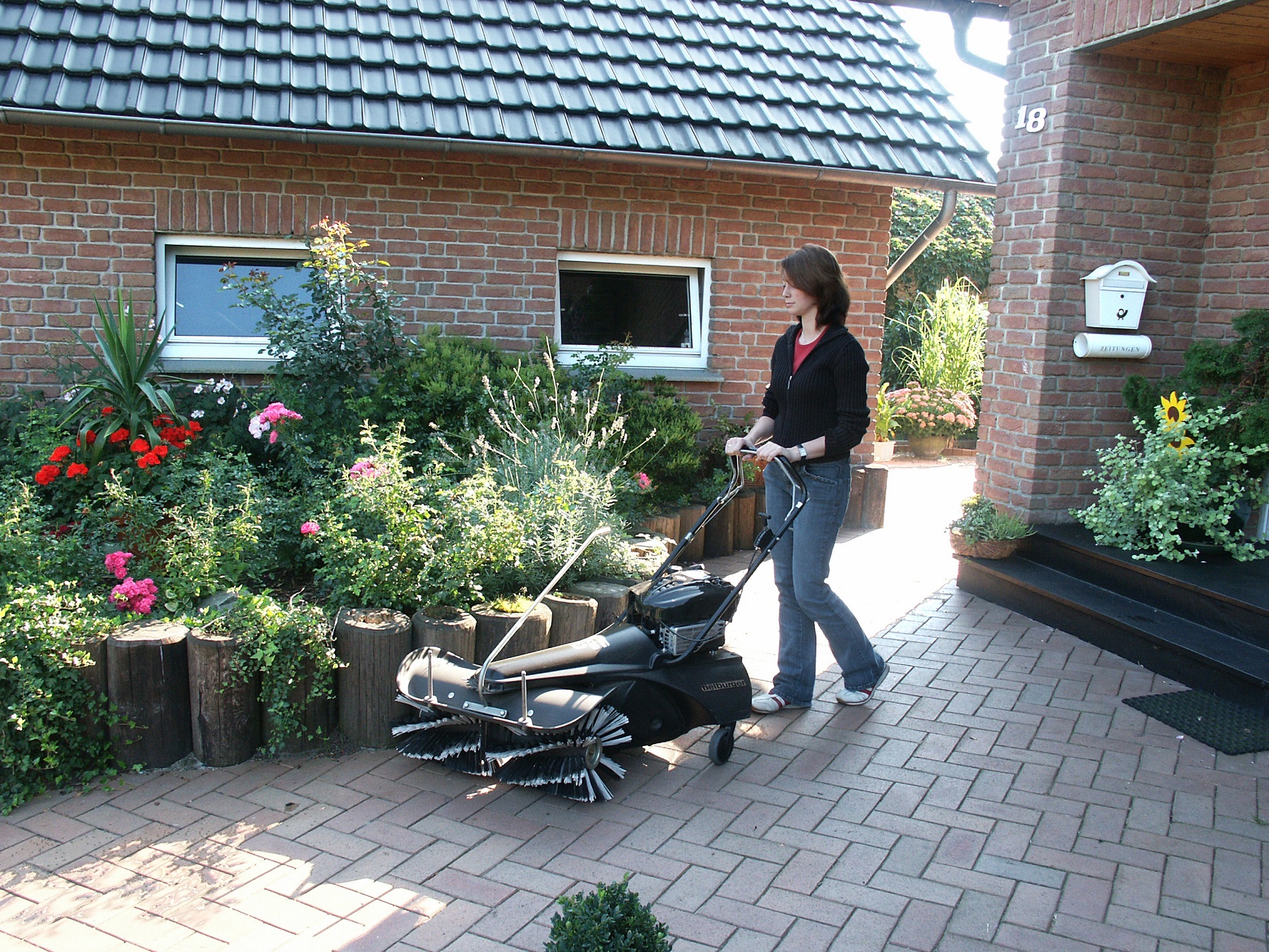 Kobieta na podwórzu domu jednorodzinnego, zamiatająca chodnik przy kwiatach za pomocą zamiatarki Tielbuerger